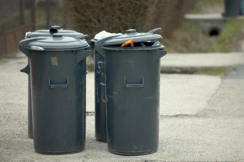 Two dark grey plastic wheeled rubbish bins with closed and partially open lids are positioned on a paved pavement in an outdoor setting, with a residential street visible in the background. The bin on the left has a visibly intact lid, while the bin on the right has its lid slightly ajar, revealing a glimpse of irregularly shaped waste and an orange handle or tool inside. The bins are situated on a concrete or asphalt surface adjacent to a curb, with some greenery and trees in the distance. The overall scene suggests typical outdoor waste collection or disposal activity, consistent with rubbish removal services in the Hoxton area, with minimal environmental surroundings visible. The lighting indicates daytime, with natural light illuminating the scene clearly.