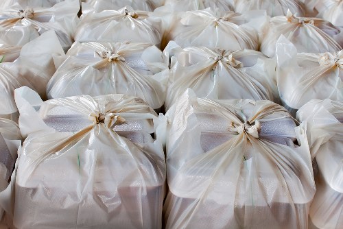 A close-up view of multiple tightly tied white plastic rubbish bags arranged in rows on a flat surface, likely on a pavement or driveway in an outdoor setting. The bags are filled with waste, with some slightly bulging and showing hints of their contents, which may include paper, plastic, or lightweight refuse. The plastic bags have a glossy finish and are secured at the top with evenly twisted knots. The background is blurred, indicating an open space possibly in an urban area. The scene suggests waste collection or disposal in progress, relevant to commercial waste removal services offered by Commercial Waste Removal Hoxton, located in the London borough area, as inferred from the postcode and local context. The lighting appears natural, emphasizing the texture and color of the bags, aligning with the professional waste management activities typical of rubbish removal efforts in Hoxton.