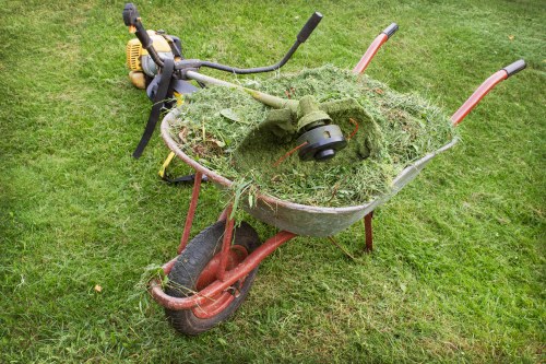 A red and black wheelbarrow positioned on a grassy outdoor area, filled with cut grass and plant debris, including blades of grass and small branches. The wheelbarrow's metal body shows signs of use, with some dirt and grass clippings adhering to its surface. In the background, part of a garden or lawn is visible, with the grass appearing well-maintained. The scene suggests yard work or garden clearance activity, which a rubbish removal service like Commercial Waste Removal Hoxton might handle for clients in the local Hoxton area. The overall lighting is natural, indicating daytime conditions, and the environment looks tidy apart from the garden waste in the wheelbarrow, ready for collection or disposal.