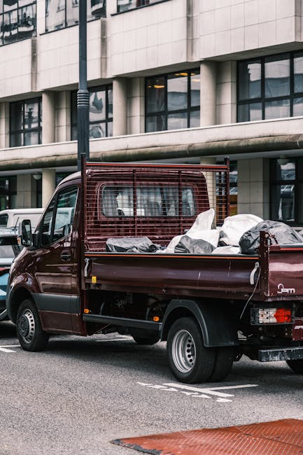 A small, dark red pickup truck with a metal mesh bumper guard attached behind the cab, parked on an urban street in front of a modern office building with large glass windows and a light-colored stone