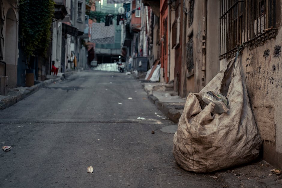 Three large black garbage bags filled with discarded waste are stacked on a paved sidewalk near a black metal fence and dense foliage. The bags appear to be made of thick plastic material and are part